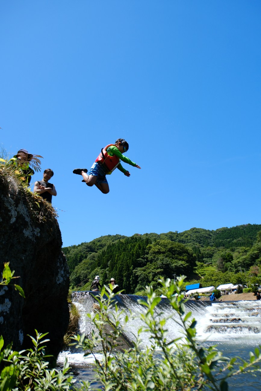天然の飛び込み台から川へ飛び込む瞬間。青空を背に宙に浮いている