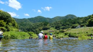 川に胸まで浸かって山をながめる親子3人の後ろ姿。青空と緑の山