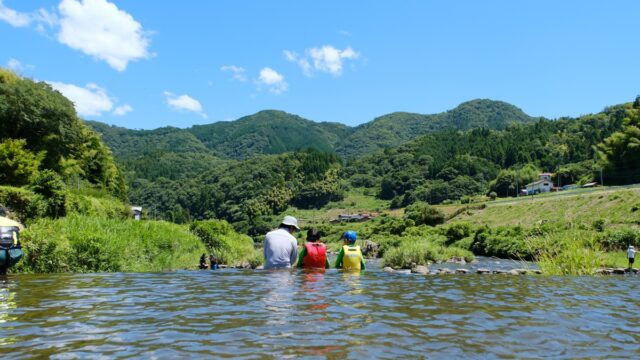川に胸まで浸かって山をながめる親子3人の後ろ姿。青空と緑の山