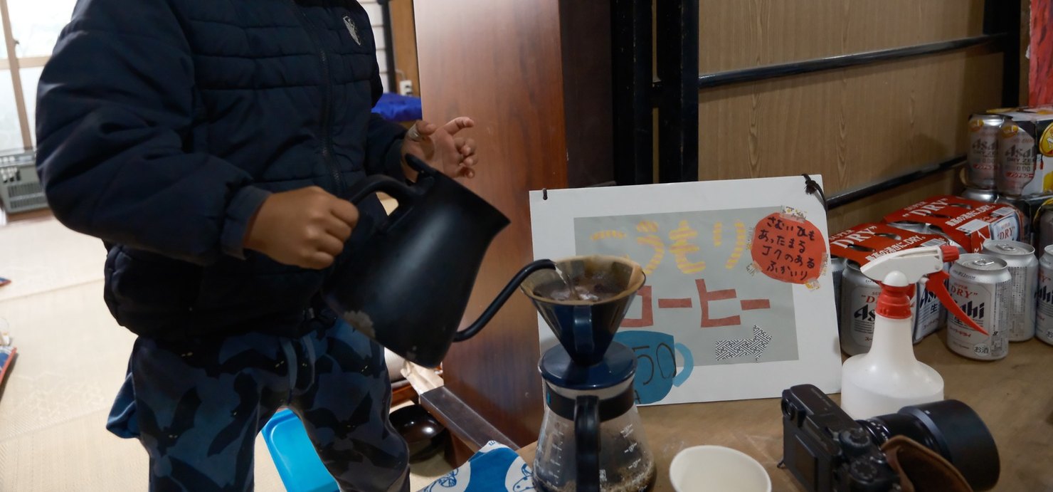 子どもがケトルでドリッパーにお湯を注いでいる。こうもりコーヒーの看板が横にある