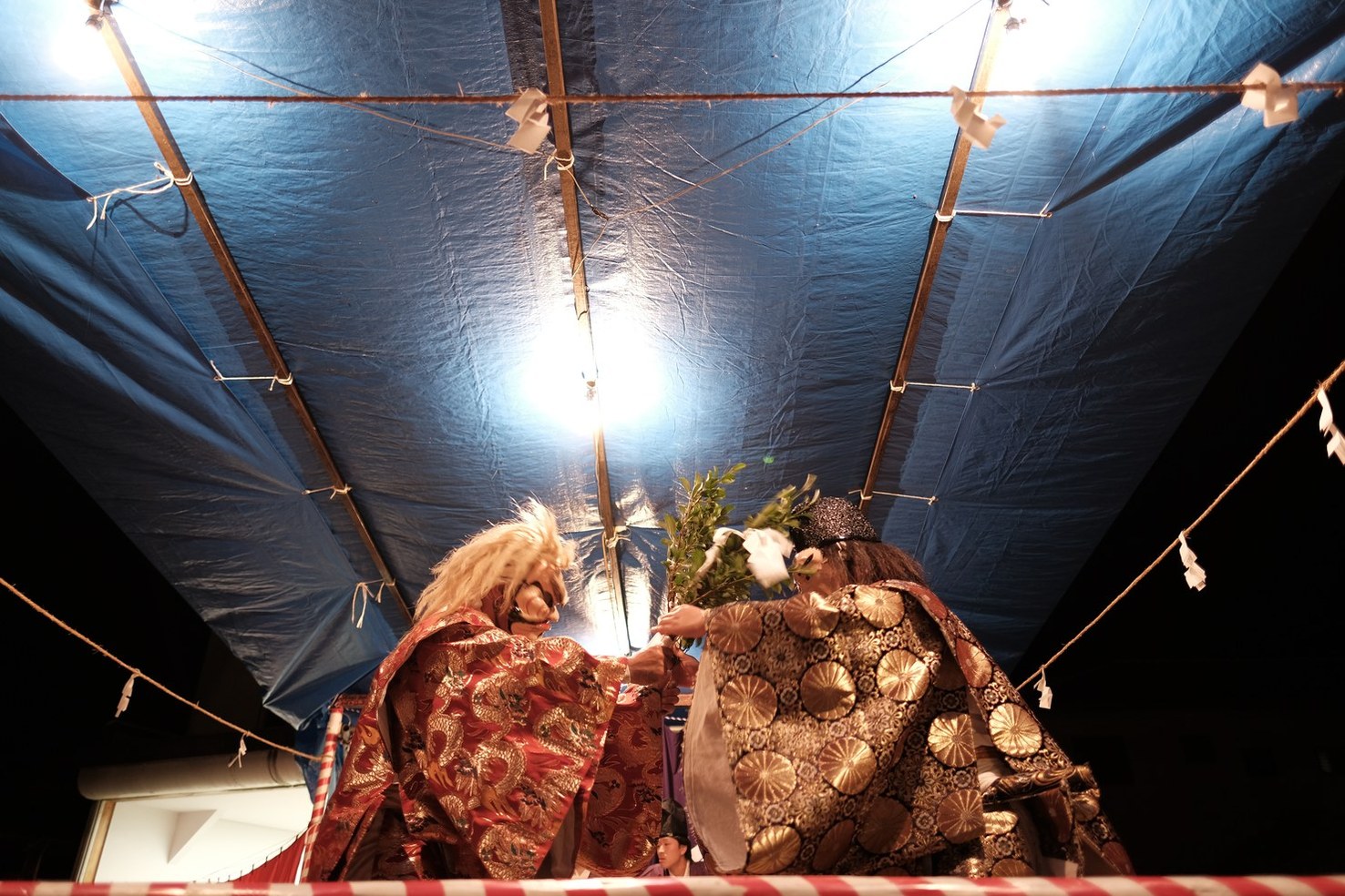 夏祭りの夜、テントの舞台で舞う神楽。二人の舞手が向かい合っている