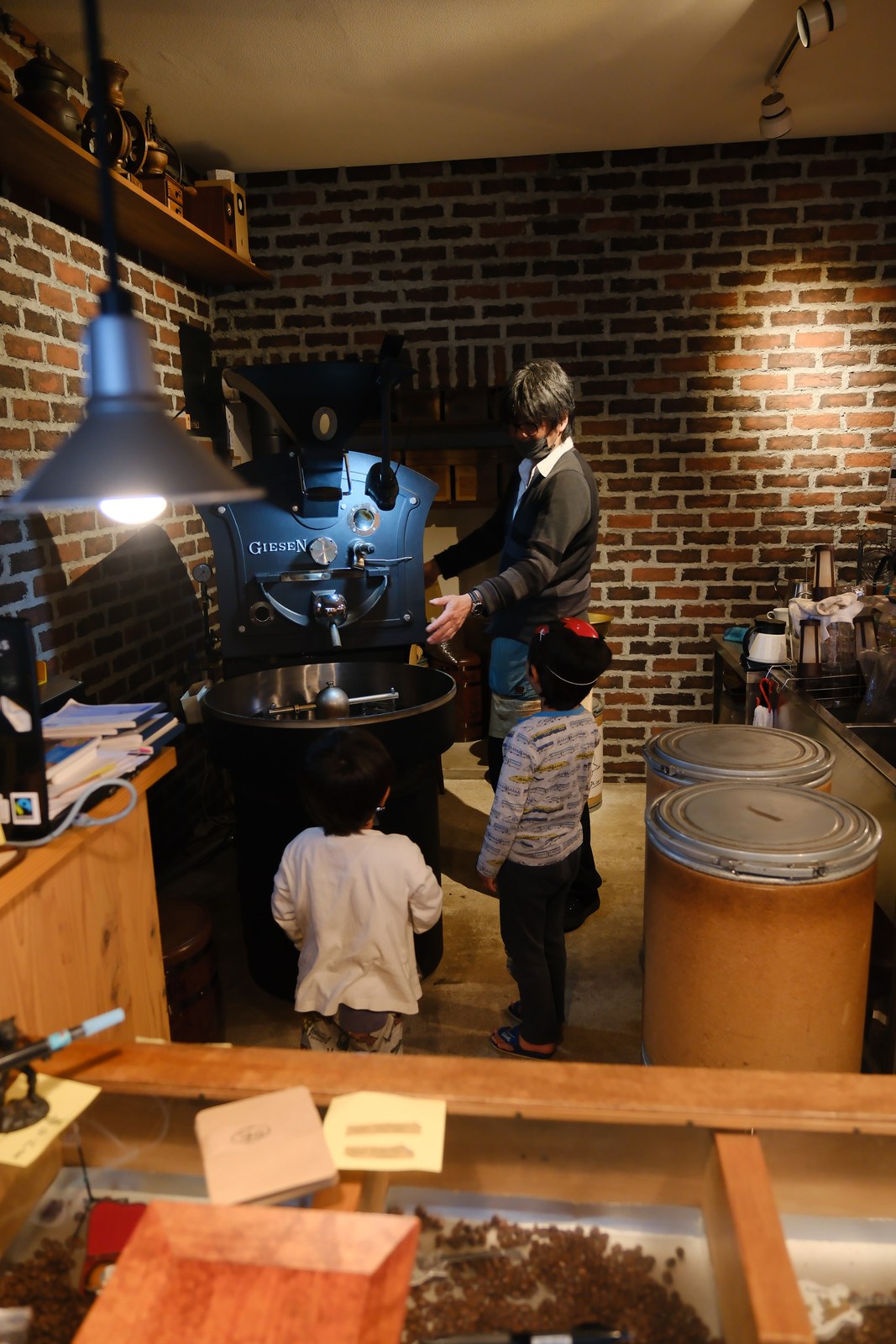 レンガの壁の前に置かれた、艶消しの黒のギーセンの焙煎機。飯島さんと、見上げる子どもたちの後ろ姿