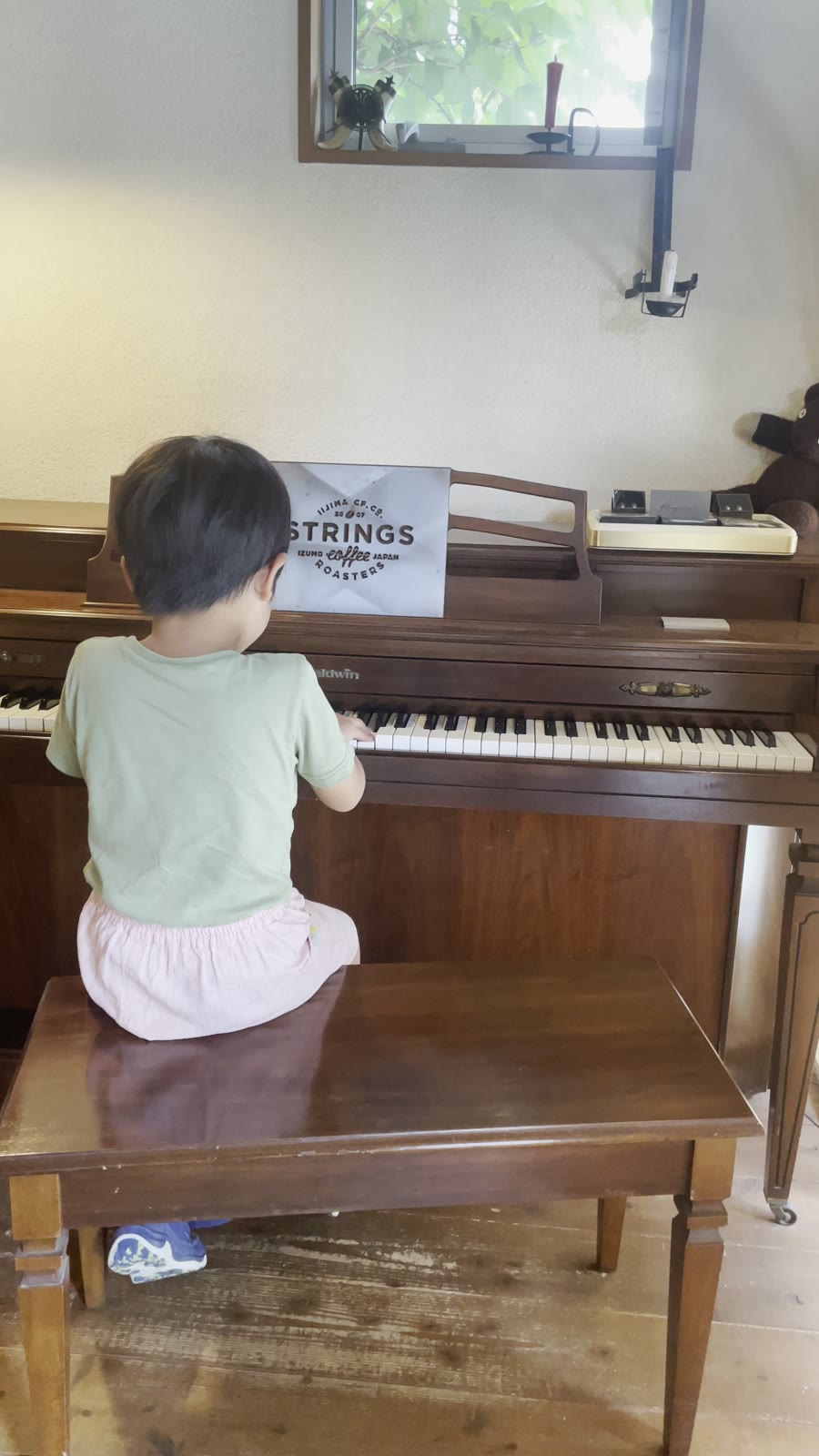 小さなアップライトピアノを弾く子どもの後ろ姿。譜面台に STRINGS COFFEE ROASTERS のロゴの札