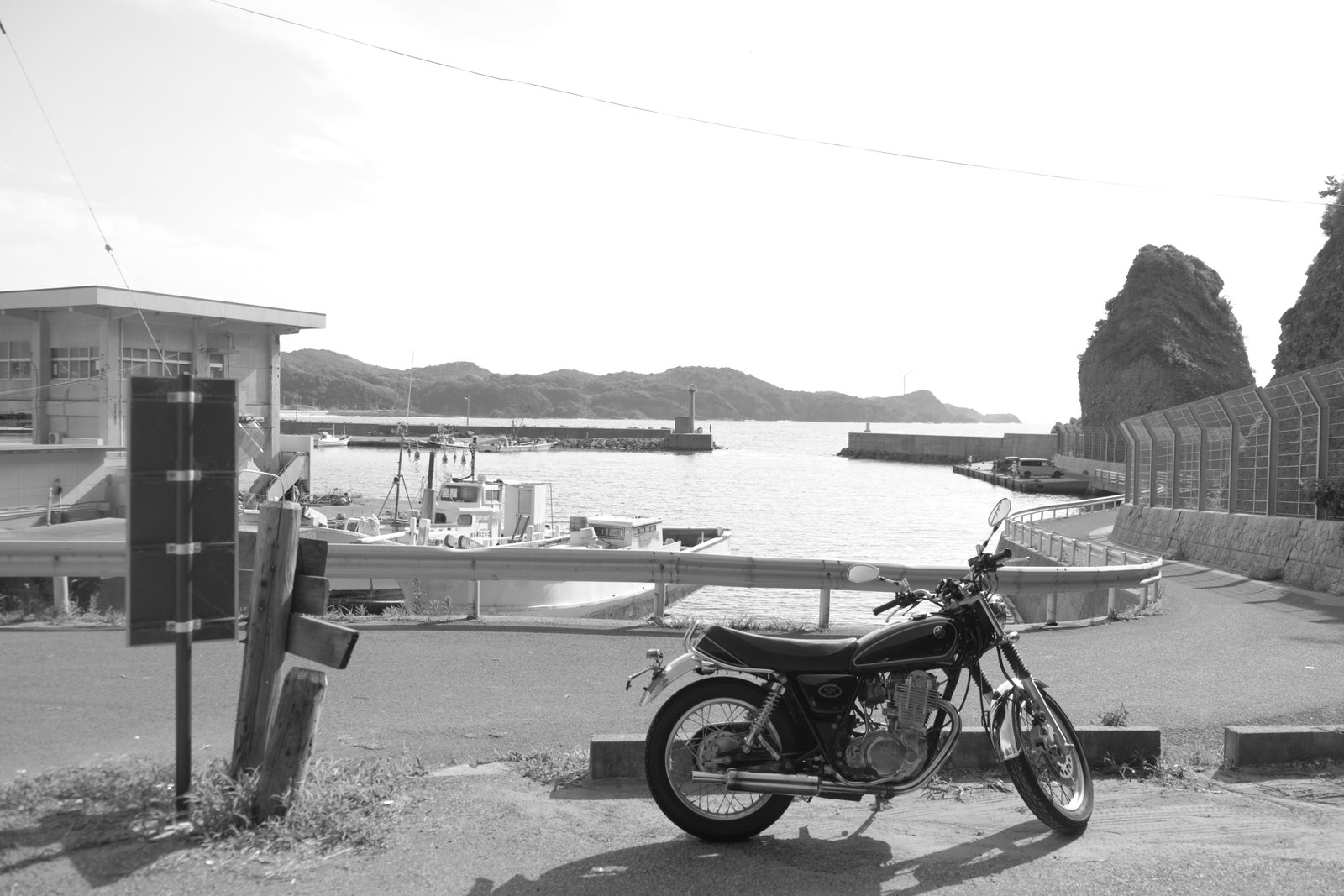 海沿いの小さな漁港の道ばたに停めた、黒いヤマハSR400。奥に漁船と防波堤、右に切り立った岩（モノクロ写真）