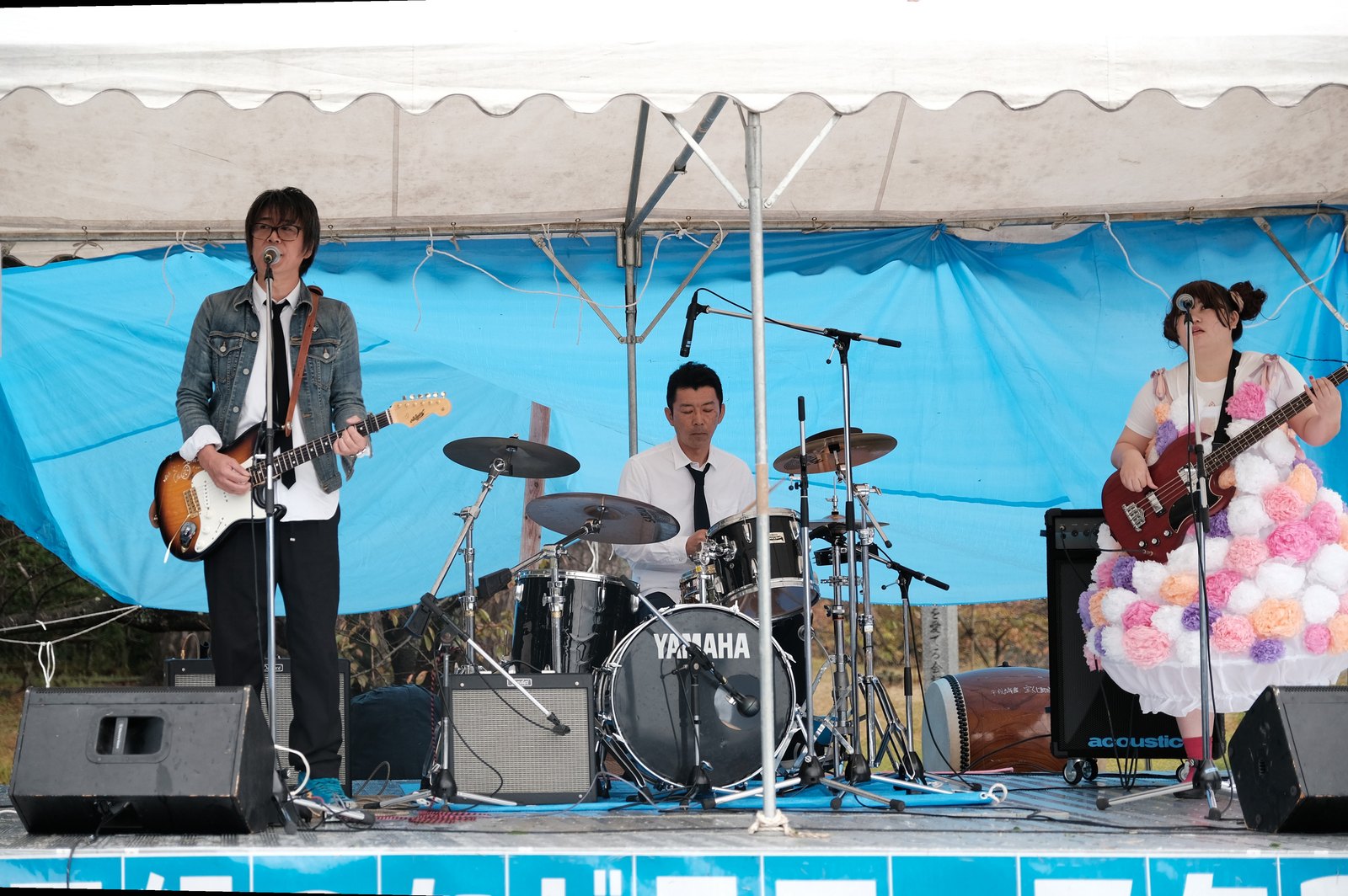 青い幕の屋外ステージで演奏するトスカペレカの3人。ギターを弾いて歌う飯島さん、ドラム、花飾りの衣装のベース
