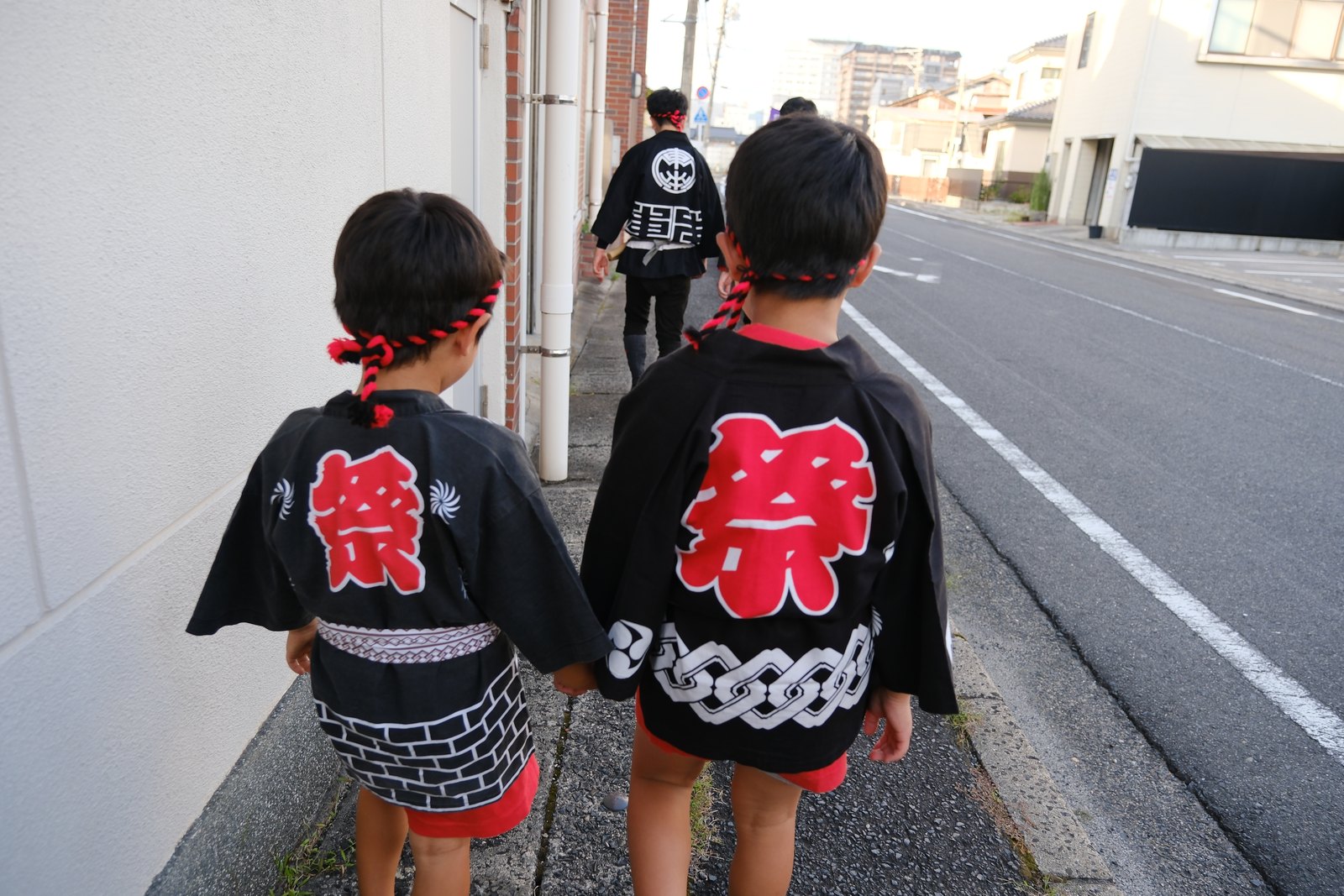 鼕行列のあと、手をつないで歩く子ども2人の後ろ姿。法被の背中に赤い「祭」の字