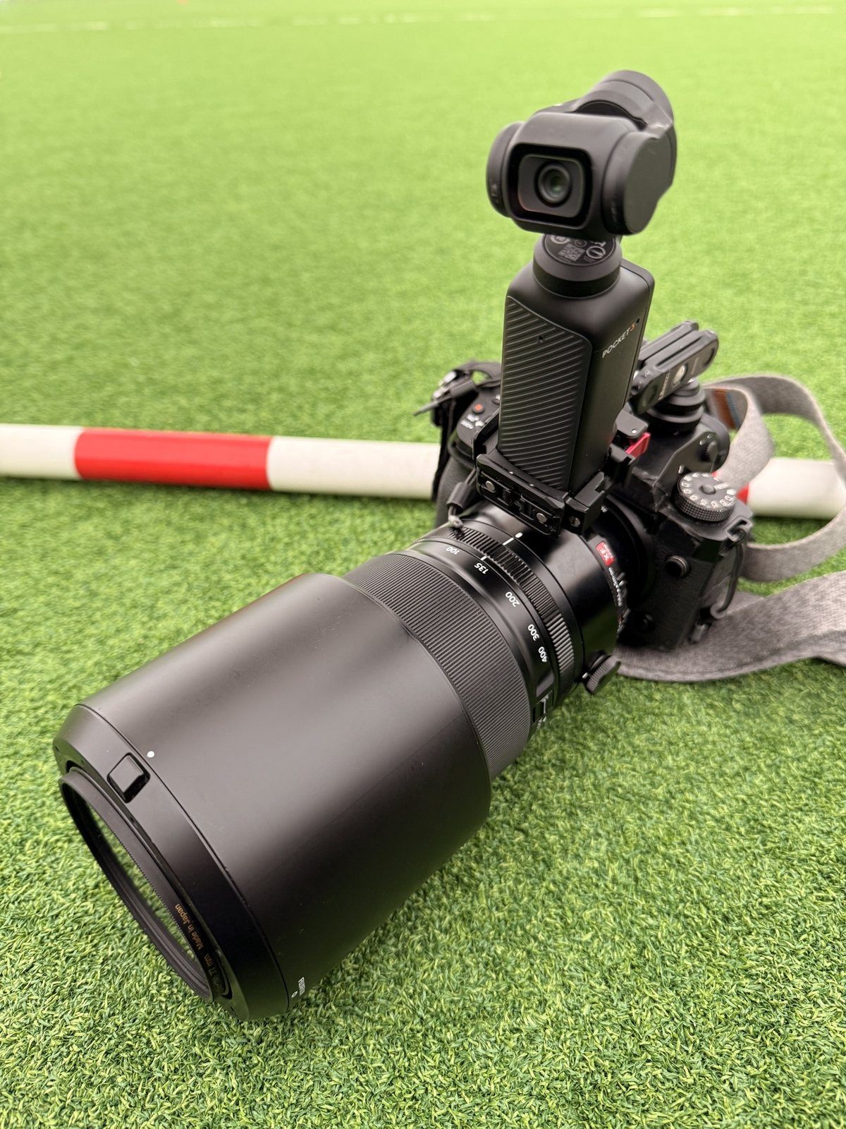 運動会の会場の芝の上に置いた一眼カメラ（望遠レンズ付き）。ホットシューのマジックアームに DJI Osmo Pocket 3 が載っている