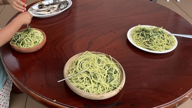 家のテラスの丸いテーブルに並んだ、ジェノヴェーゼのスパゲッティ。子どもの手がフォークで巻いている