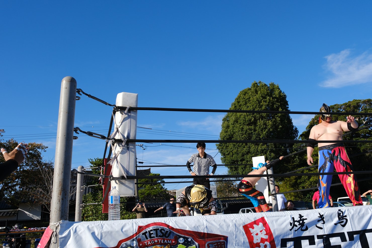 青空の下、お祭りの会場に組まれたプロレスのリング。リングの上に選手と審判