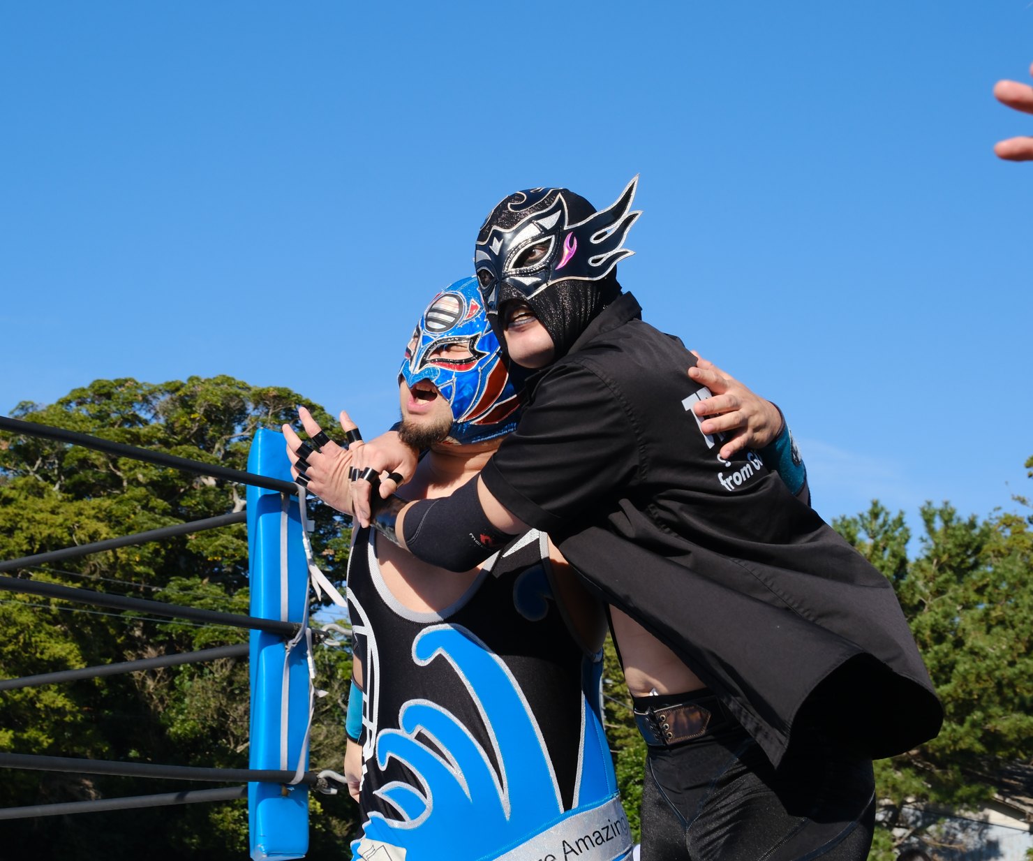 黒いマスクとコートの悪役が、青いマスクの選手をコーナーでつかまえる